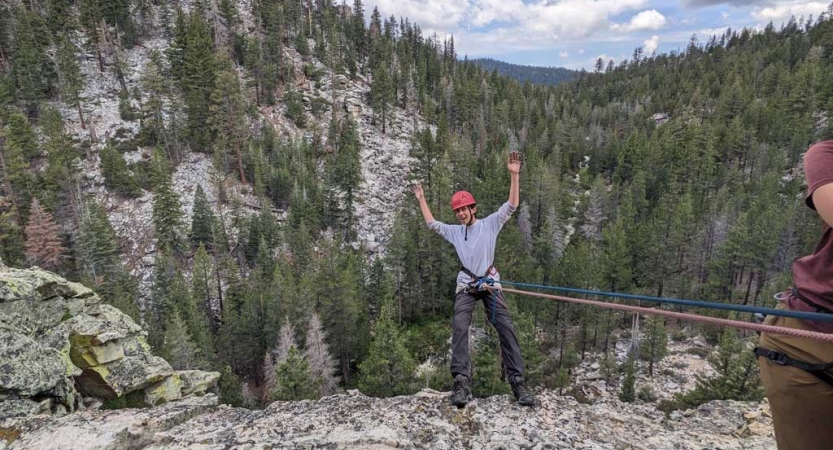 A person wearing safety gear is secured by ropes as they lean over the edge of a cliff, with trees behind and below them. 